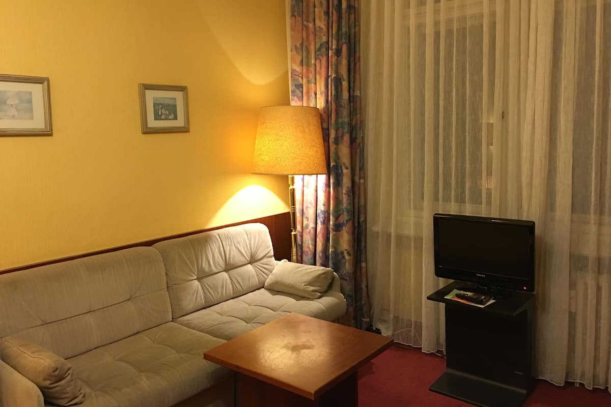 Ein Hotelzimmer mit einem beigen Sofa, einem Holztisch, einem Fernseher auf einem Schrank, einer Lampe und einem Fenster mit transparenten Vorhängen.