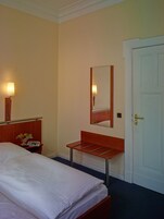 Una habitación de hotel con una cama, una mesita de noche con lámpara y frutero, un espejo y una puerta.