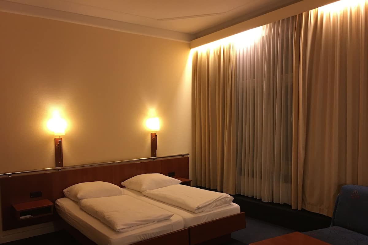 Ein Hotelzimmer mit einem Doppelbett, zwei Nachttischlampen, einem Sessel und einem Fenster mit Vorhängen.