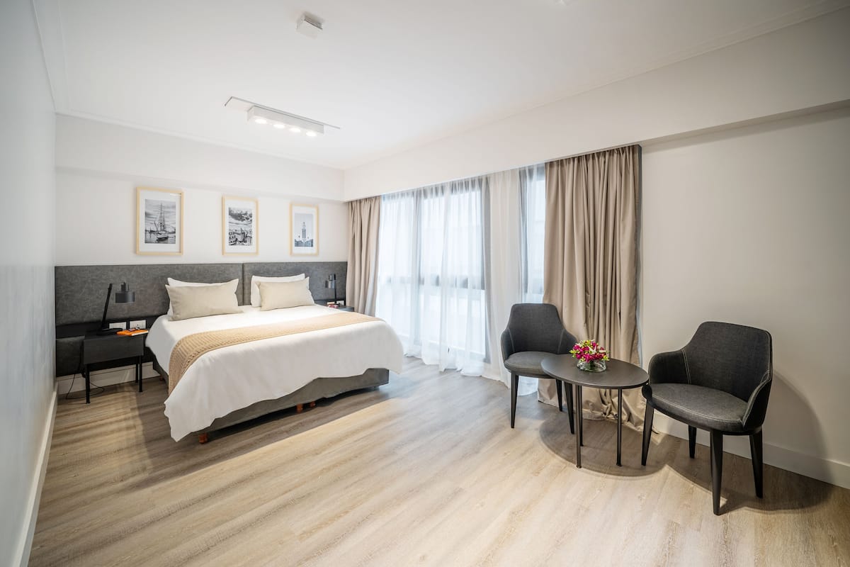 A modern hotel room with a large bed, two armchairs, a small table, and a desk with a lamp.