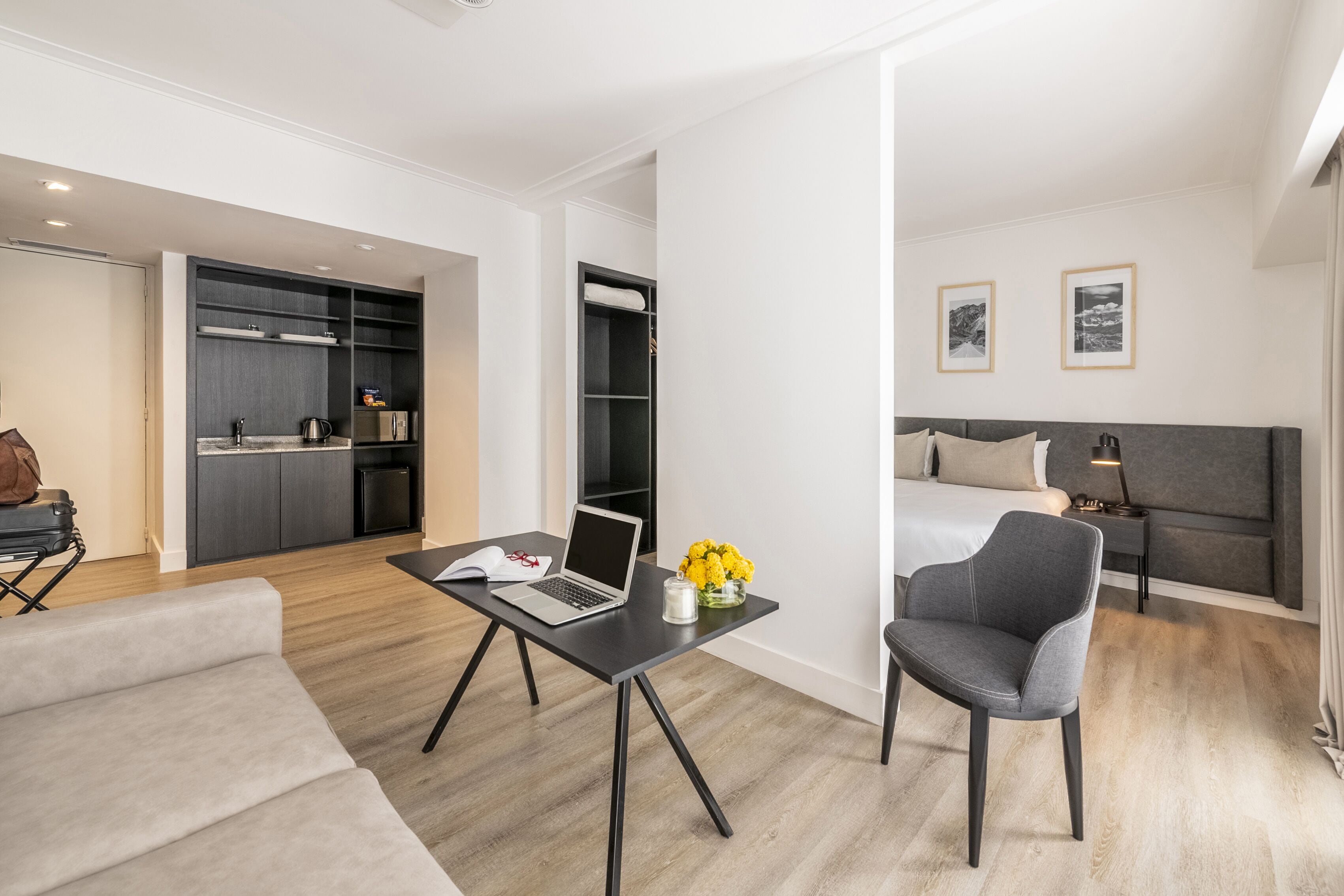 A modern hotel room with a bed, a desk with a laptop, a chair, a sofa, and a kitchenette.