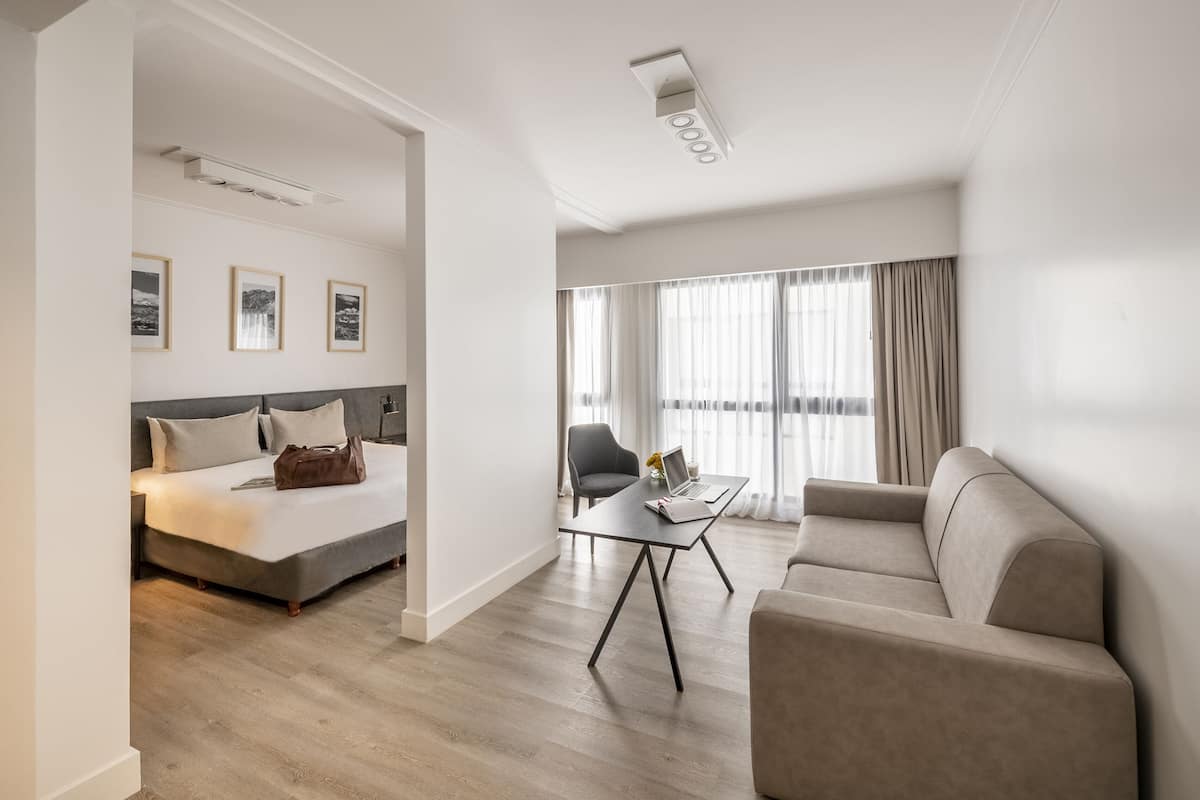 A modern hotel room with a bed, a sofa, a desk, and a large window with curtains.