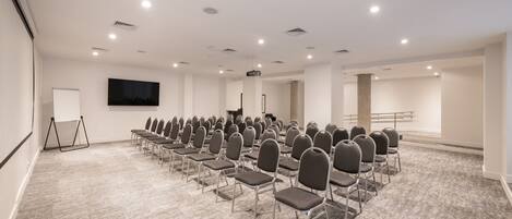 Un salón de conferencias con filas de sillas, una pantalla para proyector y una pizarra.