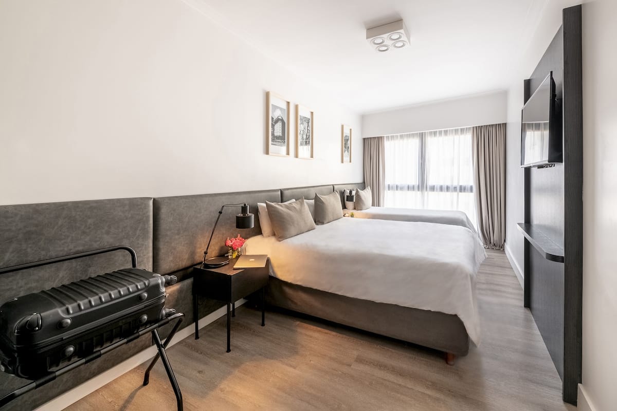 A modern hotel room with a large bed, a desk with a lamp, a suitcase, and a flat-screen TV.