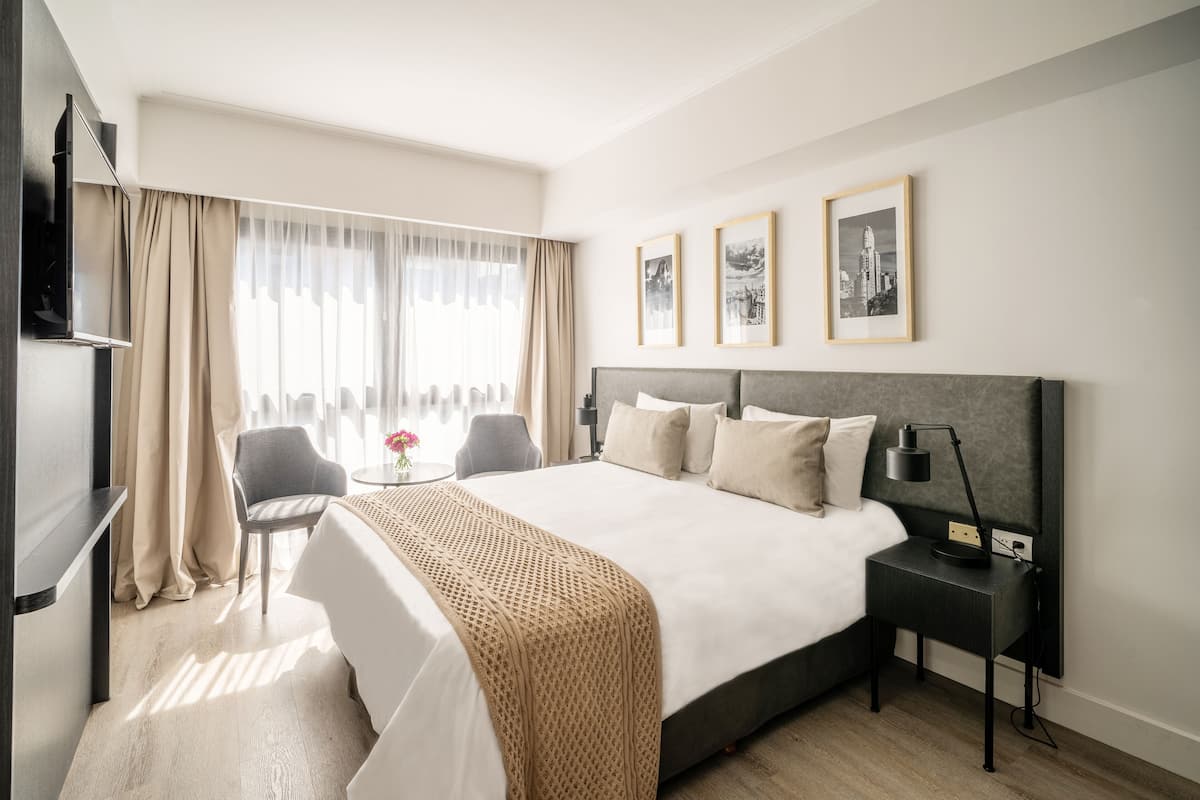 A modern hotel room with a large bed, a bedside table with a lamp, a chair, a small table with a vase, and a TV mounted on the wall.
