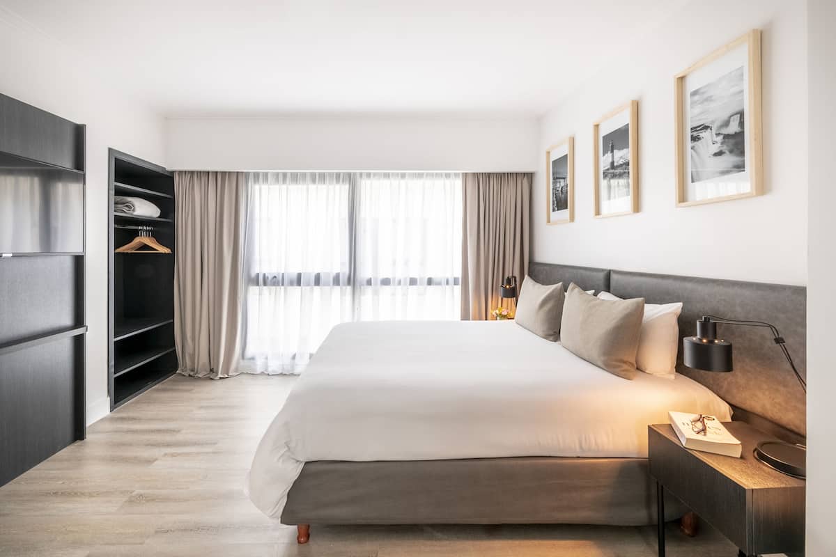 A modern hotel room with a large bed, a wardrobe, a bedside table with a lamp, and a view of the city through large windows.