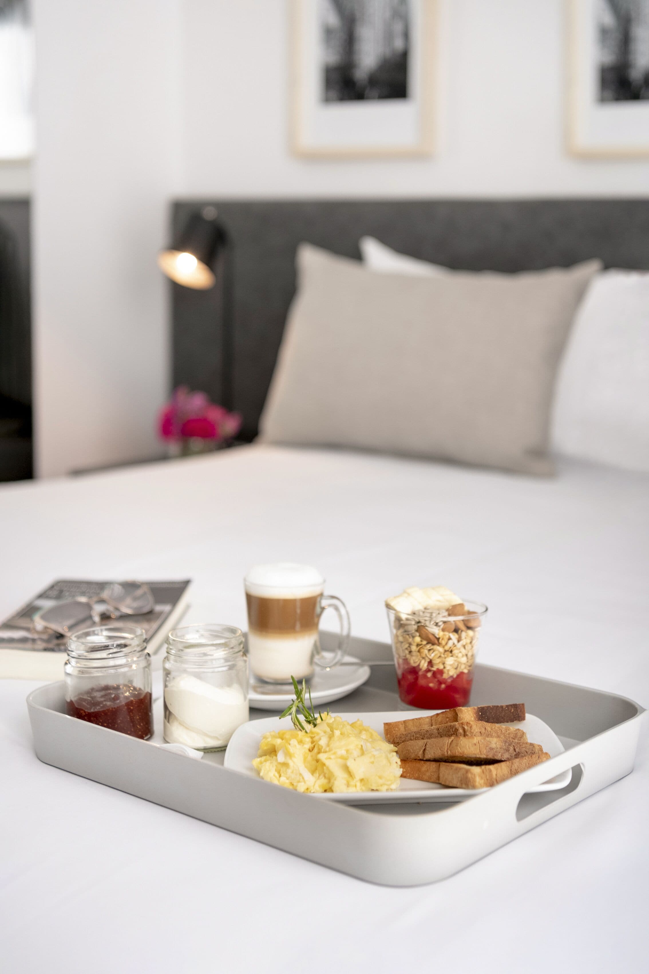 Bandeja de desayuno con tostadas, huevos revueltos, una taza de café y un vaso de leche sobre una cama en una habitación de hotel.