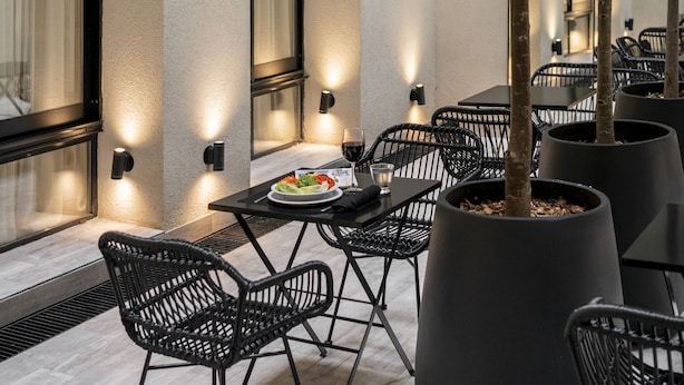 Un espacio al aire libre con mesas y sillas, una jardinera grande con un árbol, y luces fijadas a la pared.