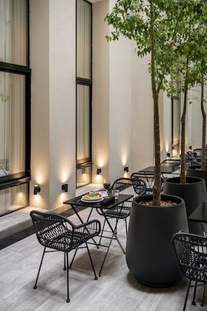 Un espacio al aire libre con mesas y sillas, una jardinera grande con un árbol, y luces fijadas a la pared.
