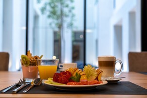 Un desayuno con un plato de fruta, un vaso de zumo de naranja y una taza de café sobre una mesa con vistas a un edificio y un árbol.