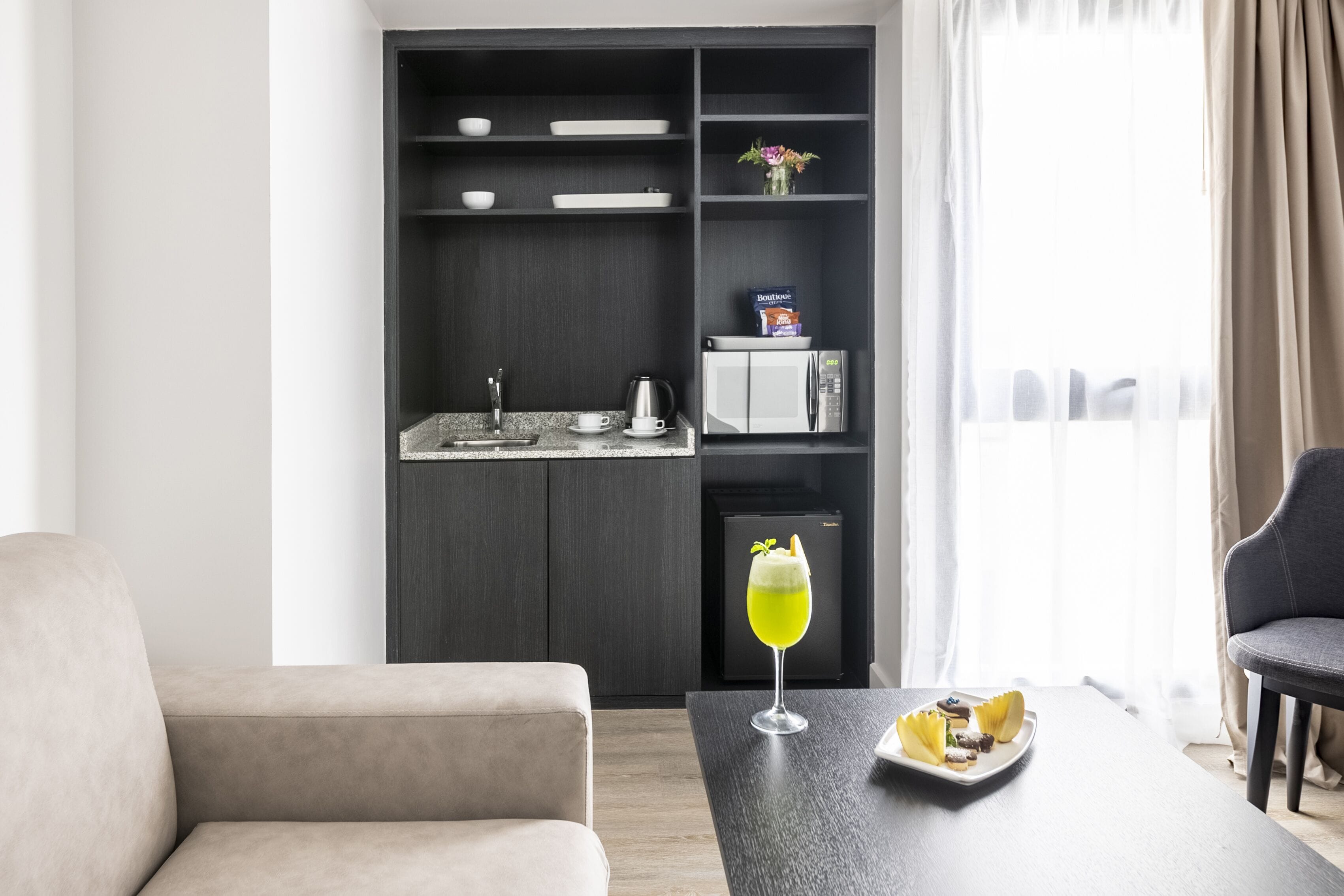 A modern living room with a sofa, a dining table with a glass of drink and snacks, a kitchen area with a fridge, microwave, and cabinets.