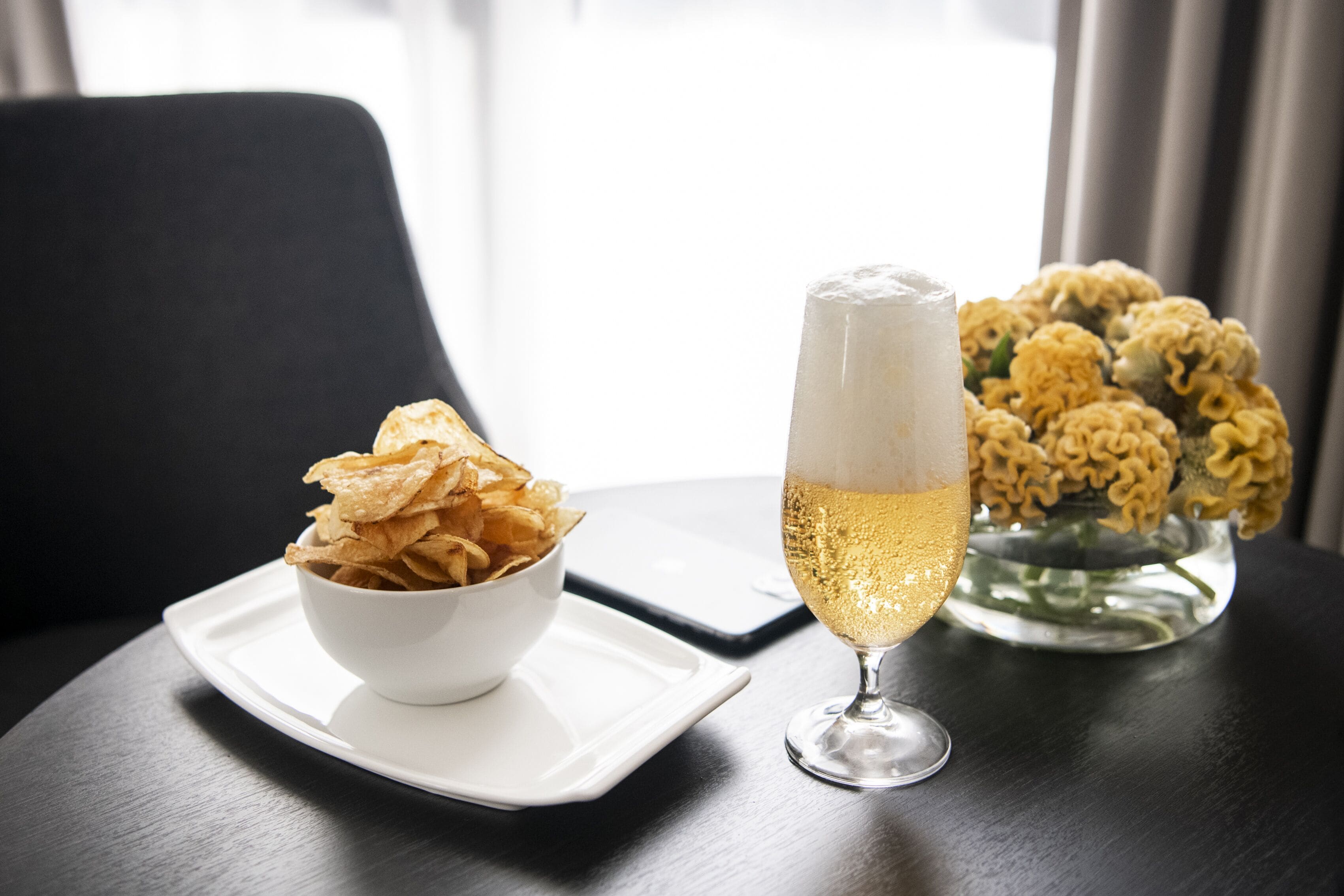 A white bowl of chips on a white plate, a glass of champagne, and a vase of yellow flowers on a dark table.