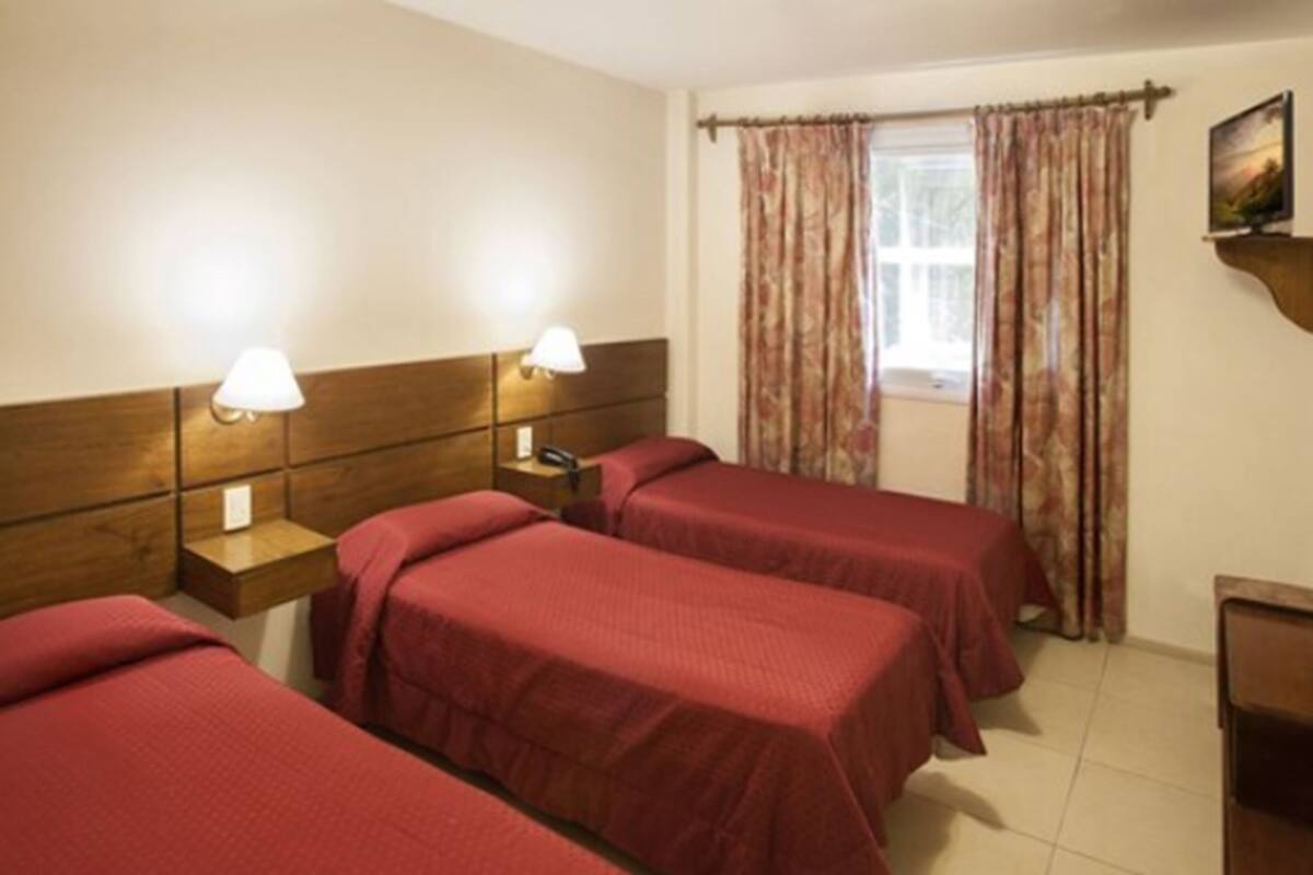 A hotel room with two beds, wooden headboards, bedside lamps, a window with curtains, and a small bedside table with a phone.
