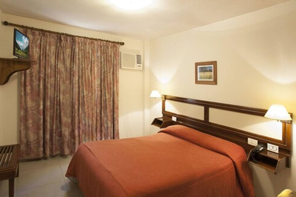 A hotel room with a bed, bedside lamps, a television on a shelf, and a window with curtains.