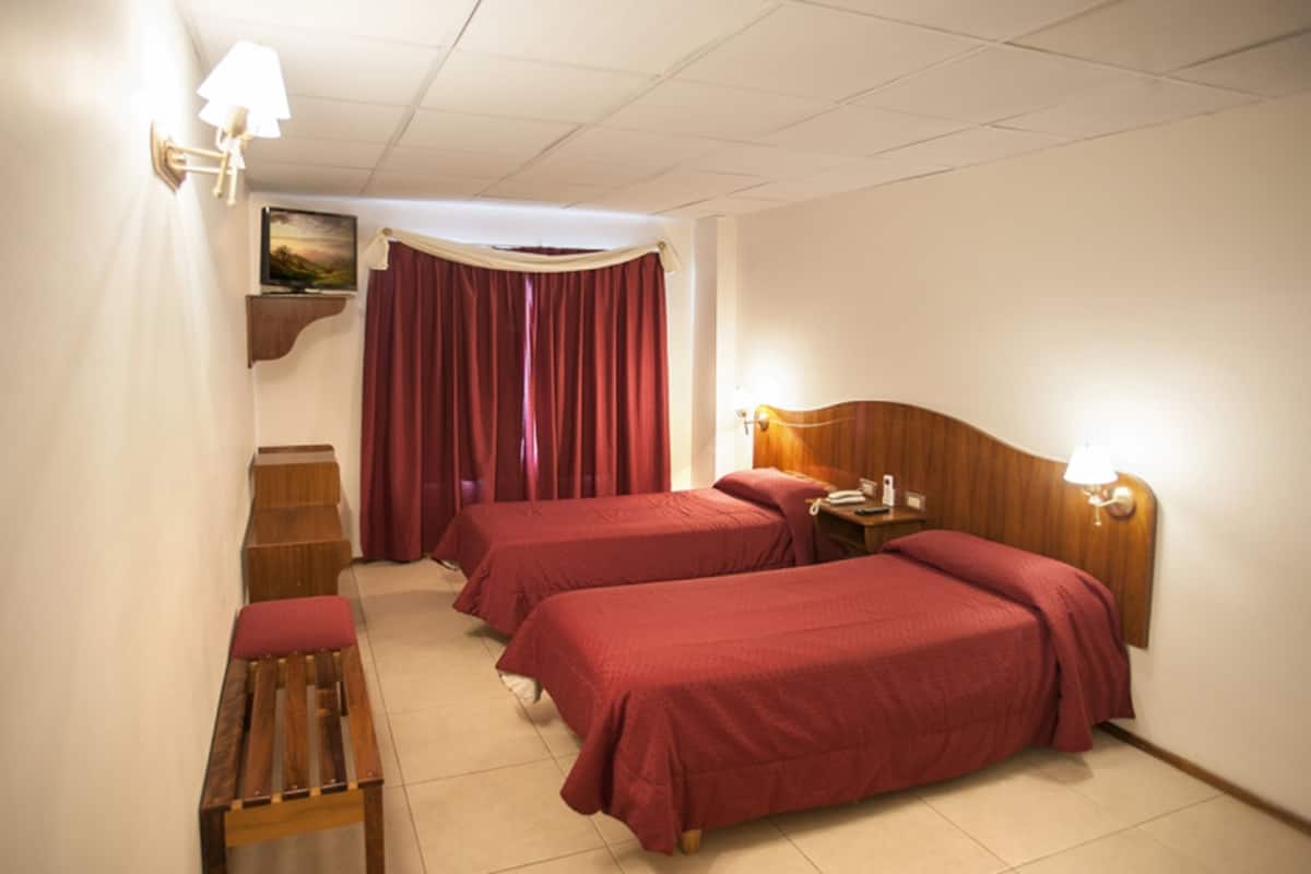 Une chambre d’hôtel avec deux lits simples, un banc en bois, une table de chevet avec une lampe et un tableau au mur.
