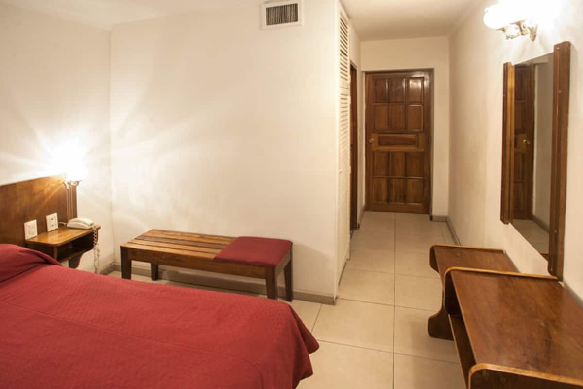 Une chambre d’hôtel avec deux lits, un banc en bois, une table de chevet avec une lampe et une porte donnant sur une autre pièce.