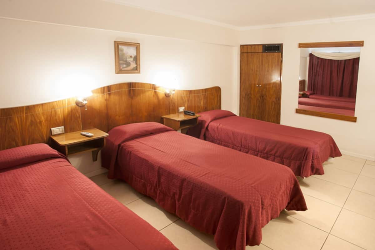 Une chambre d’hôtel avec trois lits simples, des têtes de lit en bois, des tables de chevet avec des lampes, un miroir et une armoire en bois.
