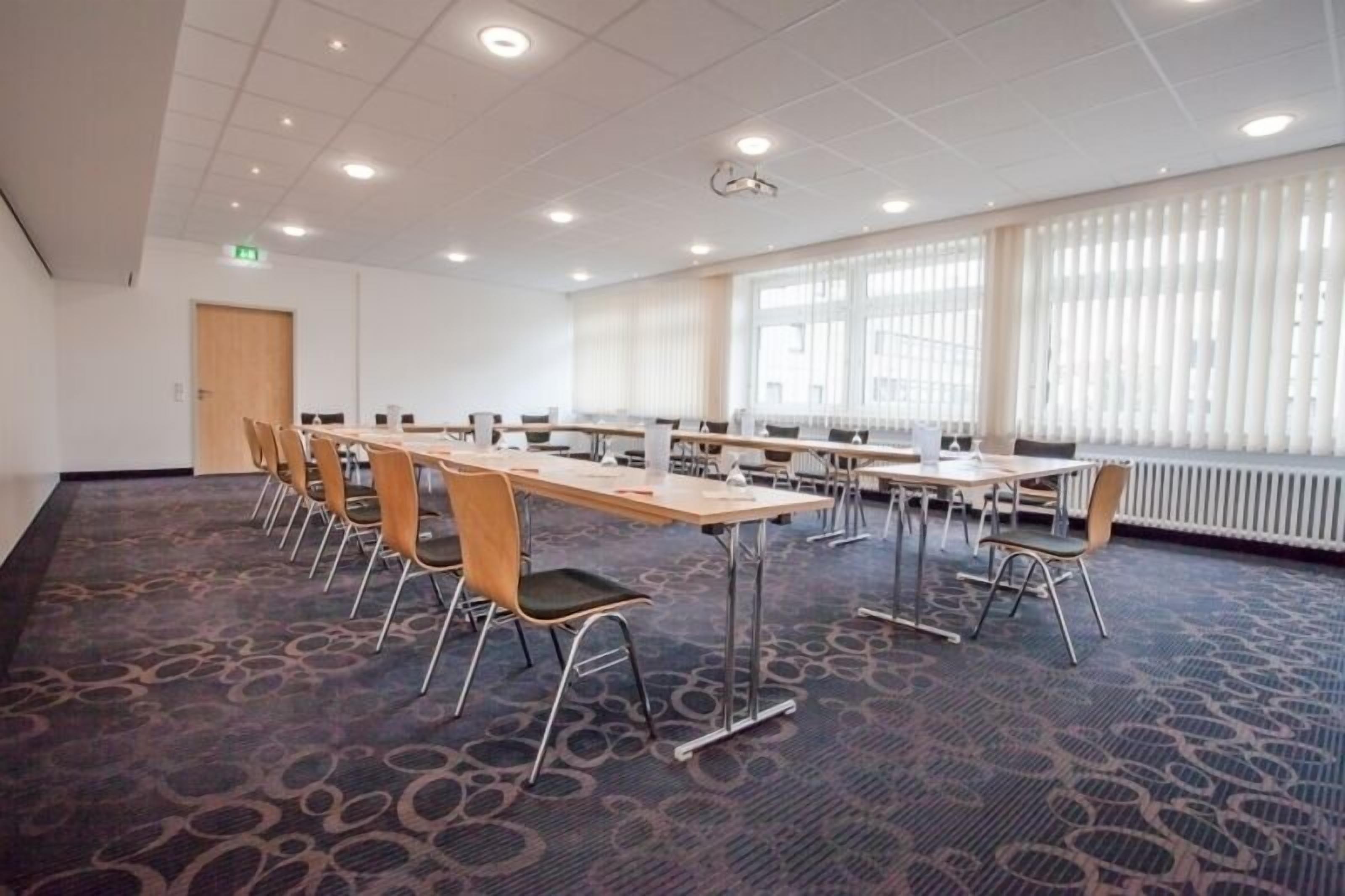 Une salle de conférence dotée de plusieurs tables et chaises, de grandes fenêtres équipées de stores et d’un sol recouvert de moquette.