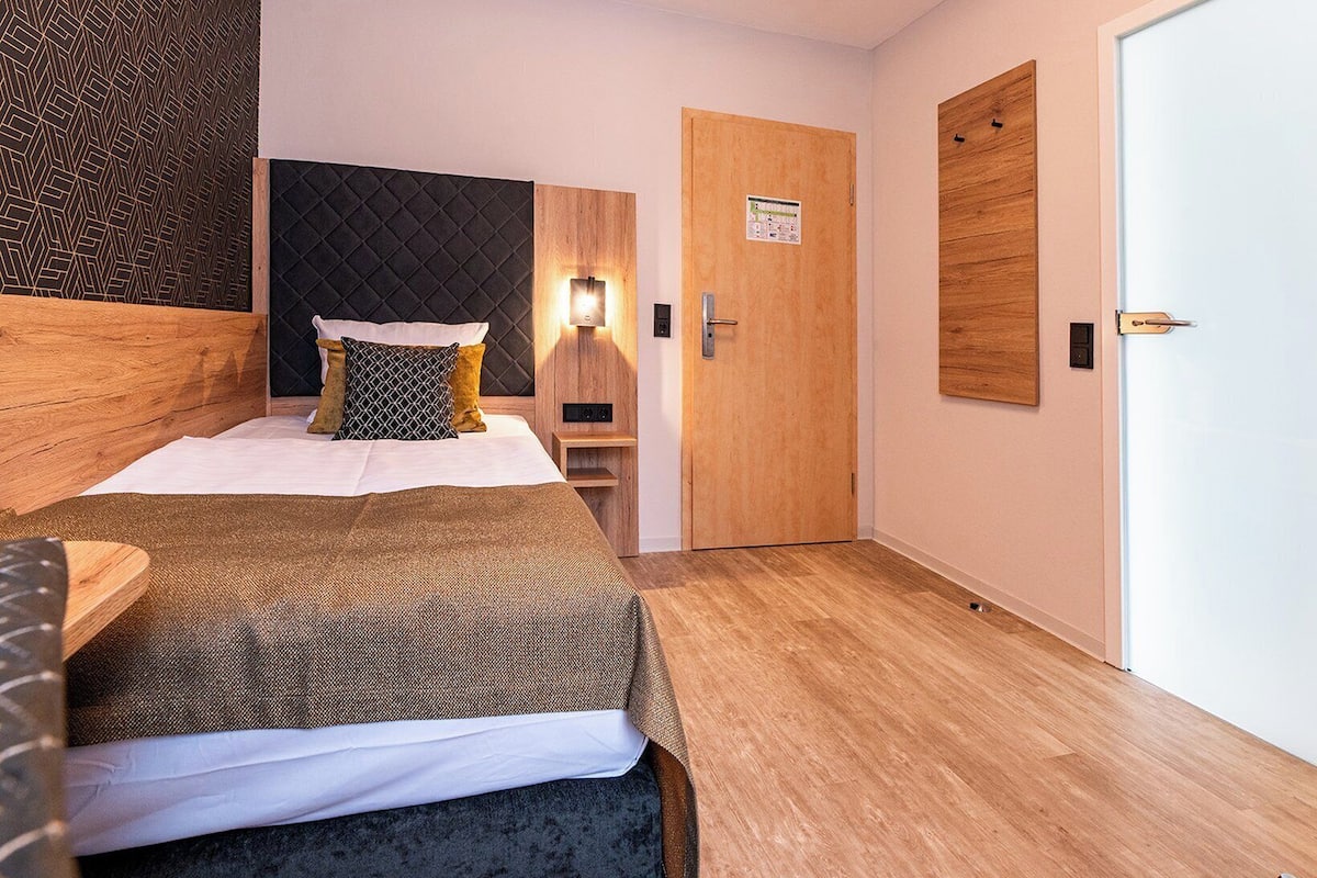 Une chambre d’hôtel moderne, équipée d’un lit, d’une table de chevet, d’une lampe fixée au mur et d’une porte en bois.