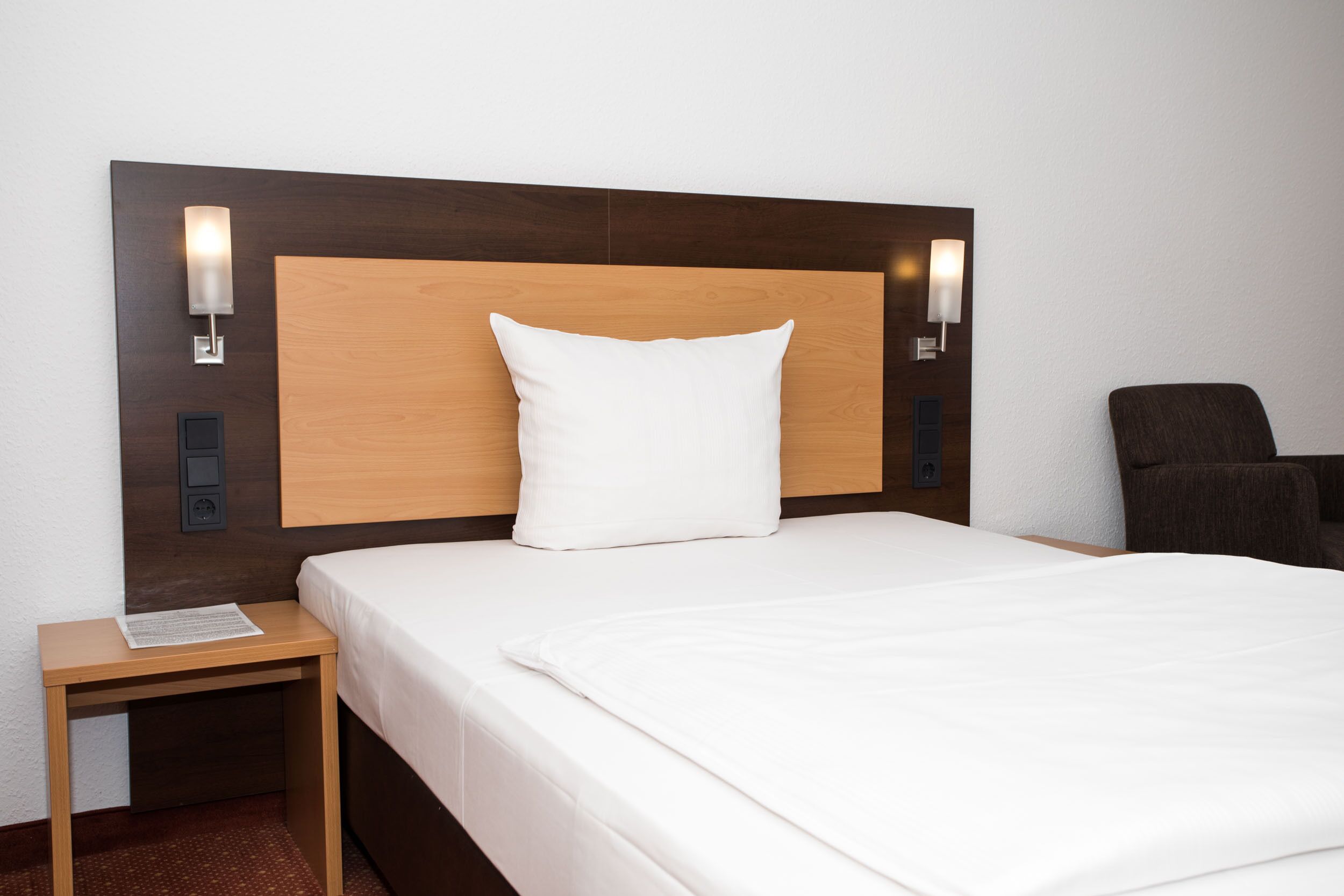 A hotel room with a wooden headboard, a white pillow, a neatly made bed, a small wooden nightstand with a lamp, and a dark armchair.