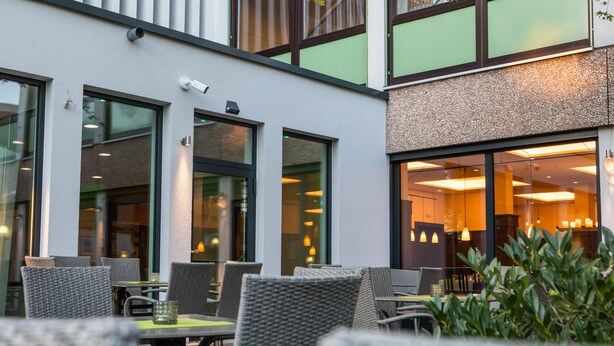 An outdoor seating area with wicker chairs and tables, large windows, and a building with a modern design. - ParkHotel Fulda (Fulda)