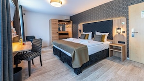 A modern hotel room with a large bed, a desk with a chair, a wardrobe, and a small bedside table.