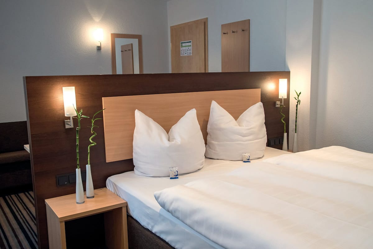 Une chambre d’hôtel avec une tête de lit en bois, deux tables de chevet avec des lampes, un lit avec une literie blanche et une petite plante sur l’une des tables.
