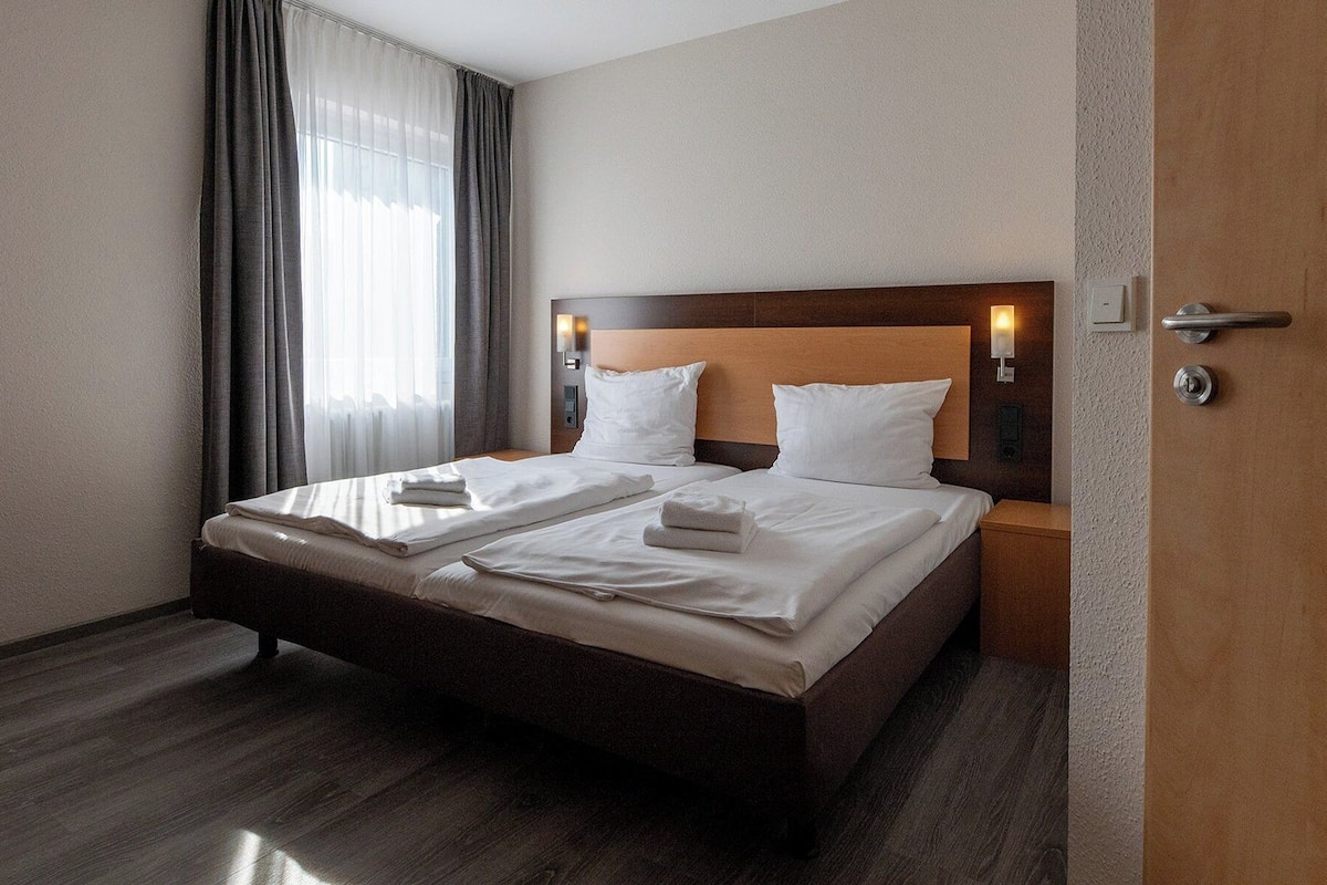 Une chambre d’hôtel avec un grand lit, deux tables de chevet, une tête de lit en bois et une fenêtre avec des rideaux.