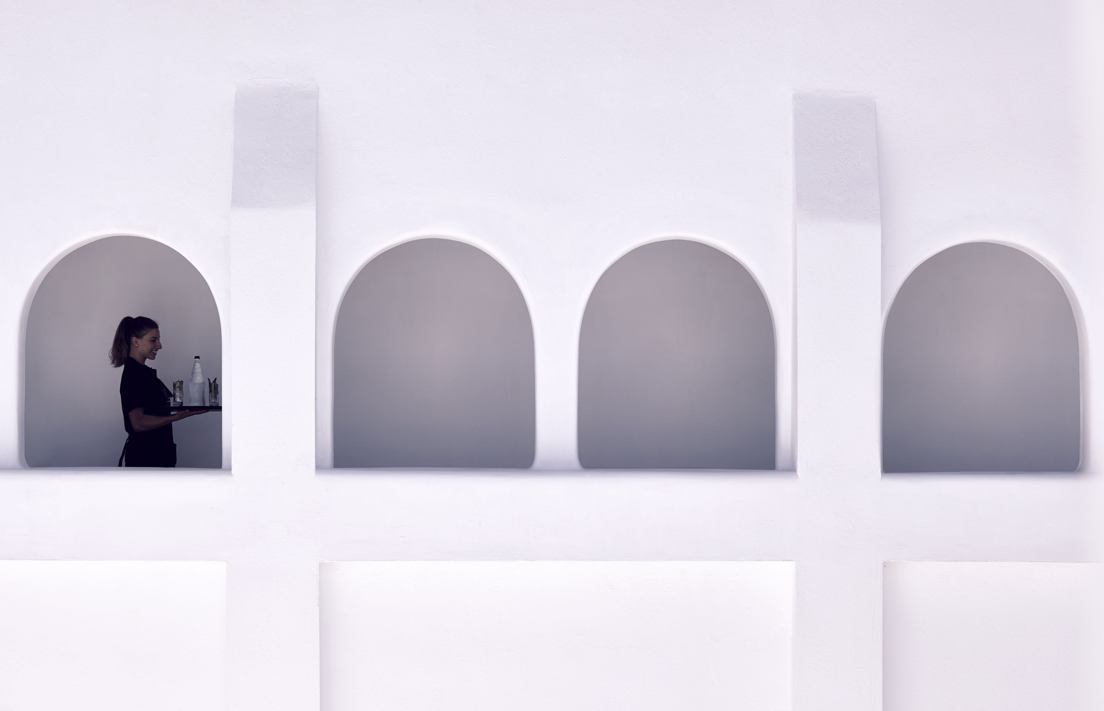 A person carrying a tray with items in a modern, minimalist room with white walls and arched niches.