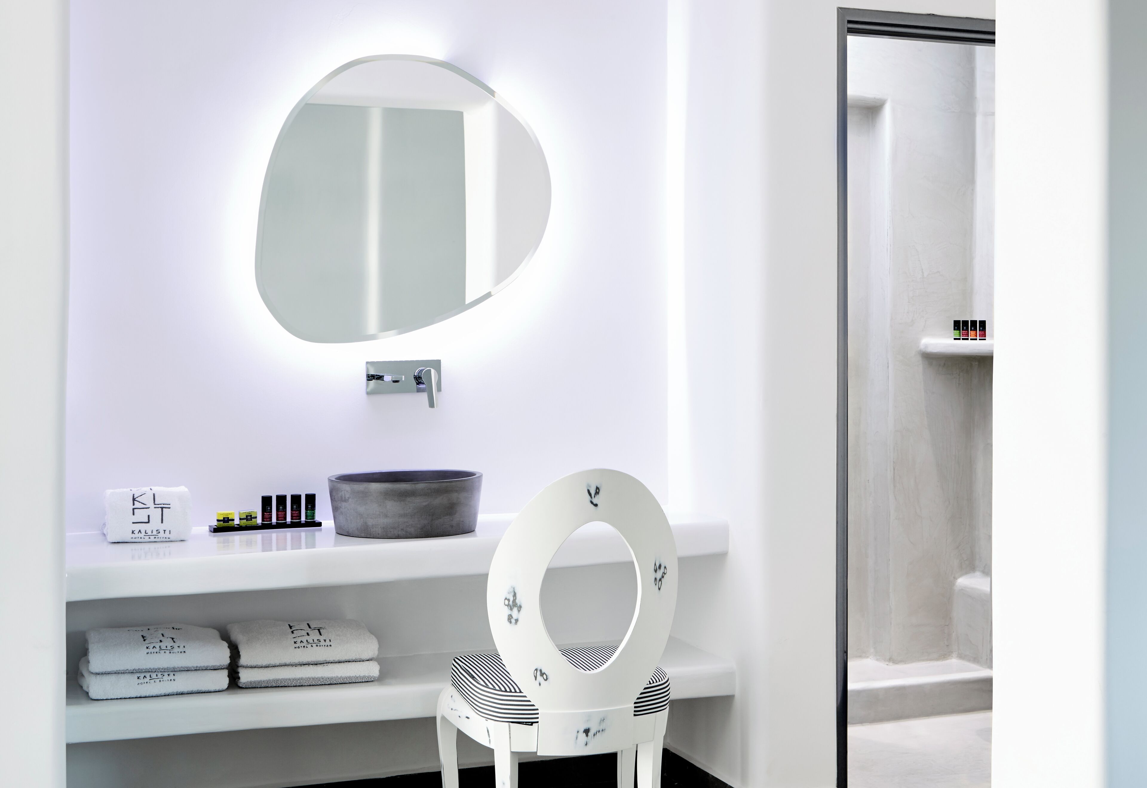 A bathroom with a white vanity, a round mirror, and a white chair.