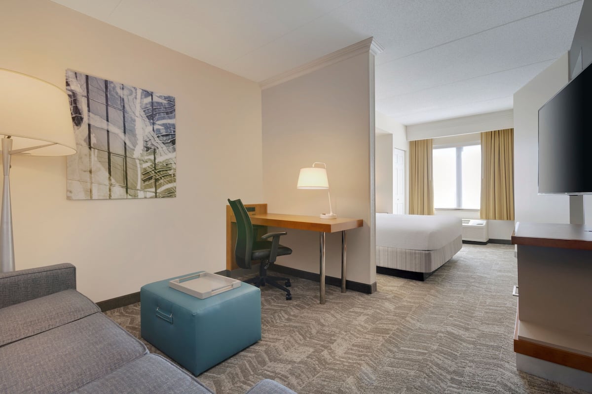 A hotel room with a sofa, a desk, a chair, a bed, a TV, and a painting on the wall.