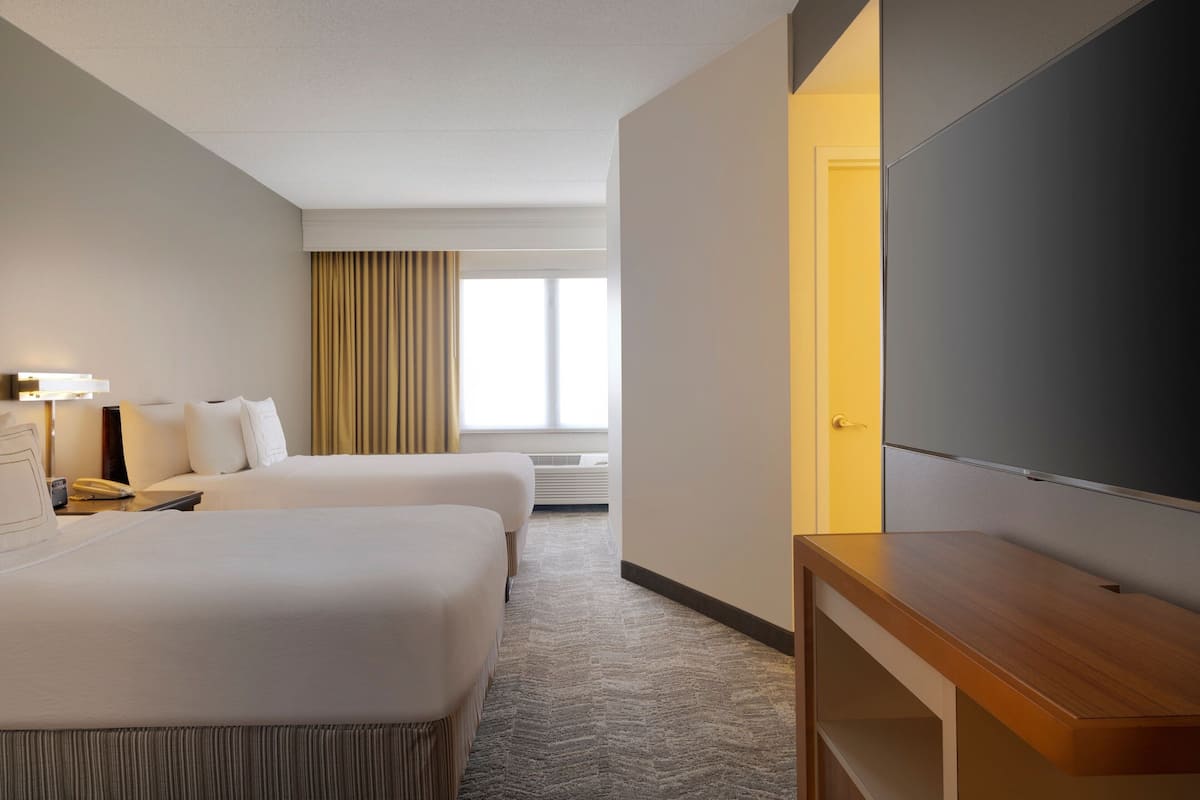A hotel room with two beds, a desk, a flat-screen TV, and a large window with curtains.