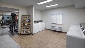 A laundry room with a row of washing machines, a dryer, a bookshelf with books, and exercise equipment. - Candlewood Suites St Robert by IHG (St. Robert)