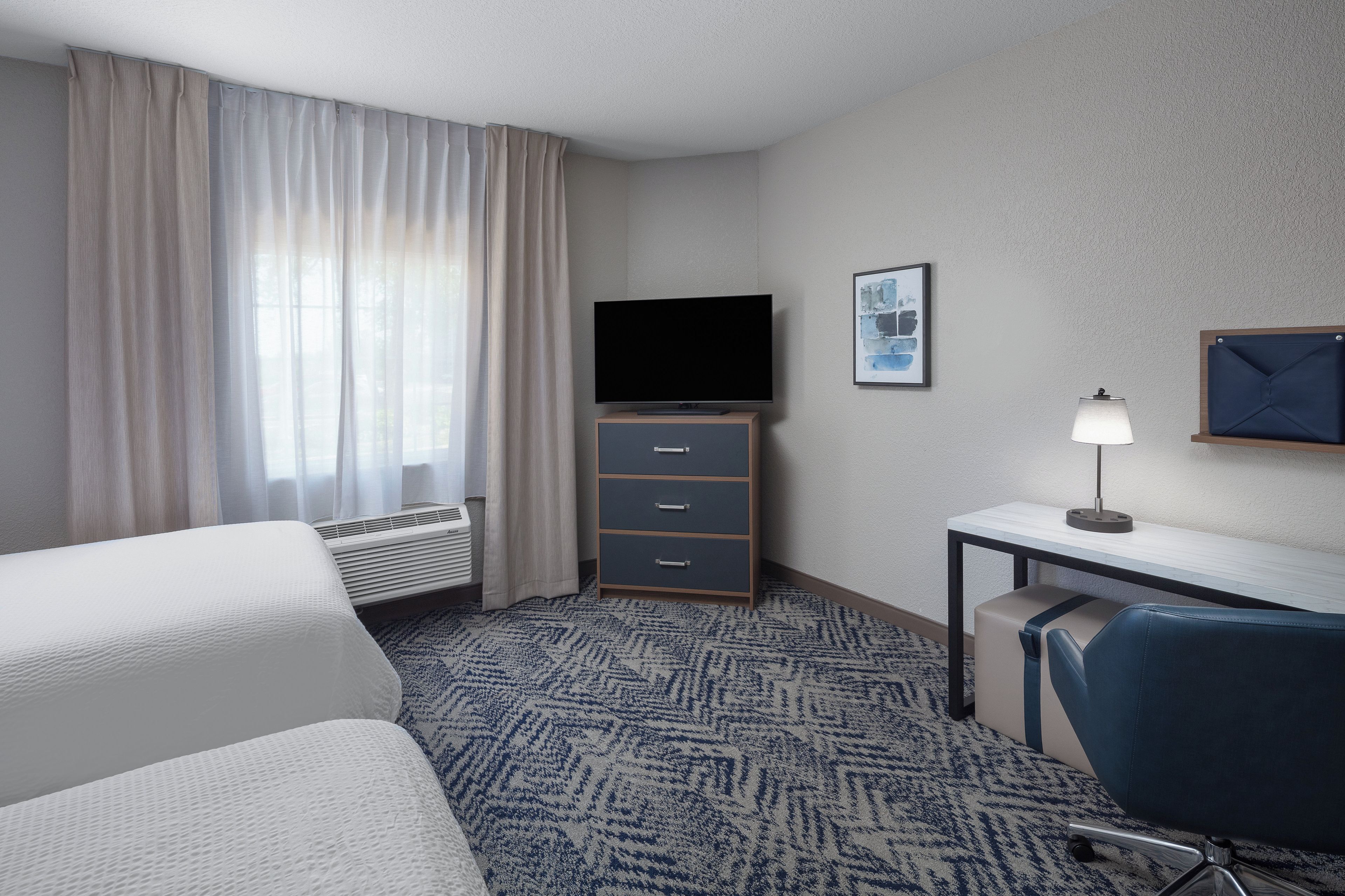 A hotel room with a bed, a desk with a lamp, a flat-screen TV, a small cabinet, and a patterned carpet.