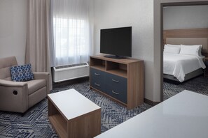 A hotel room with a bed, a TV on a wooden stand, a sofa with a patterned pillow, a coffee table, and a carpeted floor. - Candlewood Suites St Robert by IHG (St. Robert)