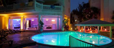 A pool area with a lounge chair, a covered seating area, and a building with balconies and a poolside bar.