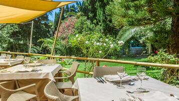 Un área de comedor al aire libre con mesas preparadas para dos personas, rodeada de abundante vegetación y una valla de bambú.