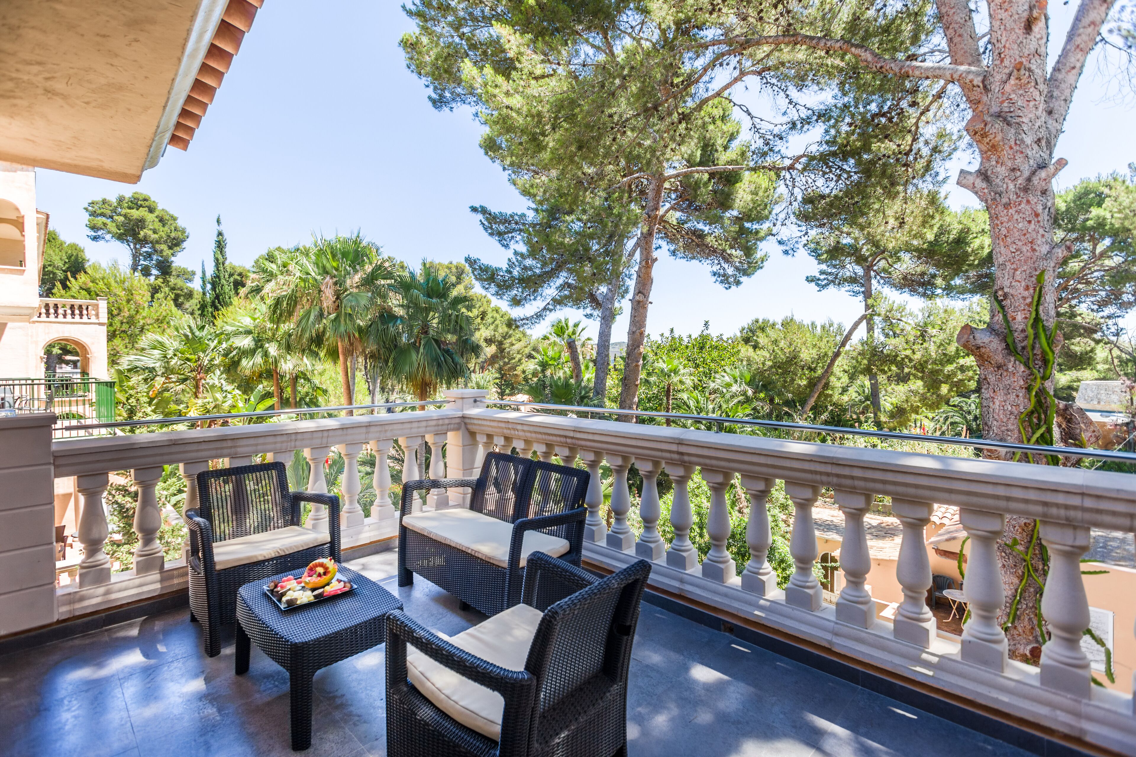 Un balcón con mobiliario de mimbre y una mesa, con vistas a un jardín con árboles y un edificio al fondo.