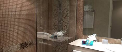 A modern bathroom with a glass-enclosed shower, a marble-tiled wall, a large mirror, and a vanity with toiletries.