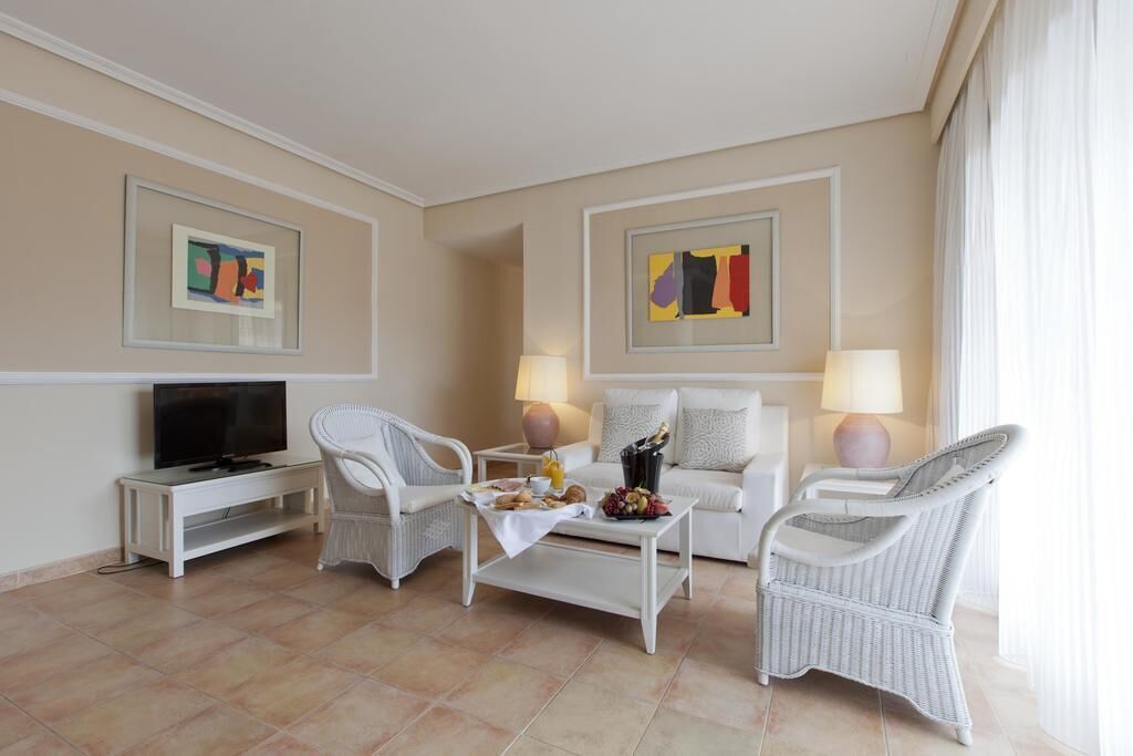 Habitación de hotel con televisión, sillas de mimbre blanco, un sofá blanco, una mesa de centro con aperitivos y obras de arte en las paredes.