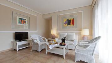 Habitación de hotel con televisión, sillas de mimbre blanco, un sofá blanco, una mesa de centro con aperitivos y obras de arte en las paredes.