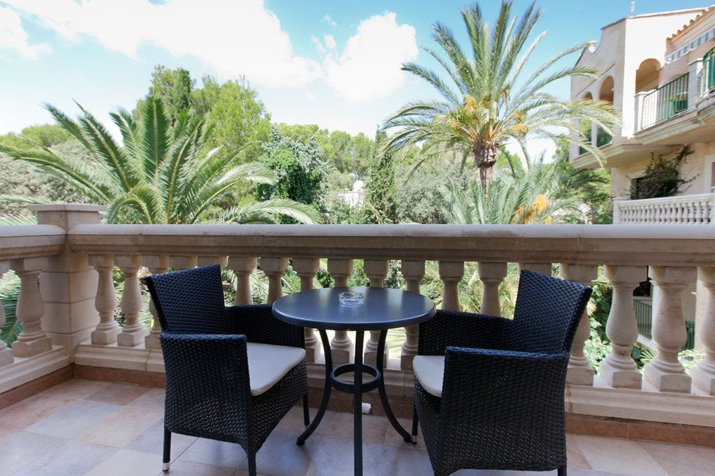 A balcony with a table and two chairs, surrounded by lush greenery and a building with classical architecture.
