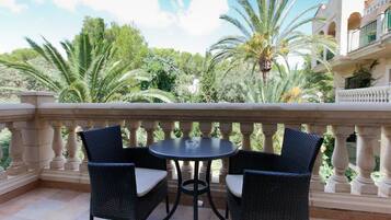 A balcony with a table and two chairs, surrounded by lush greenery and a building with classical architecture.