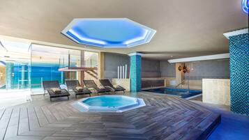 Uma área interna de piscina moderna com luminária hexagonal no teto, cadeiras de descanso e uma banheira de hidromassagem.