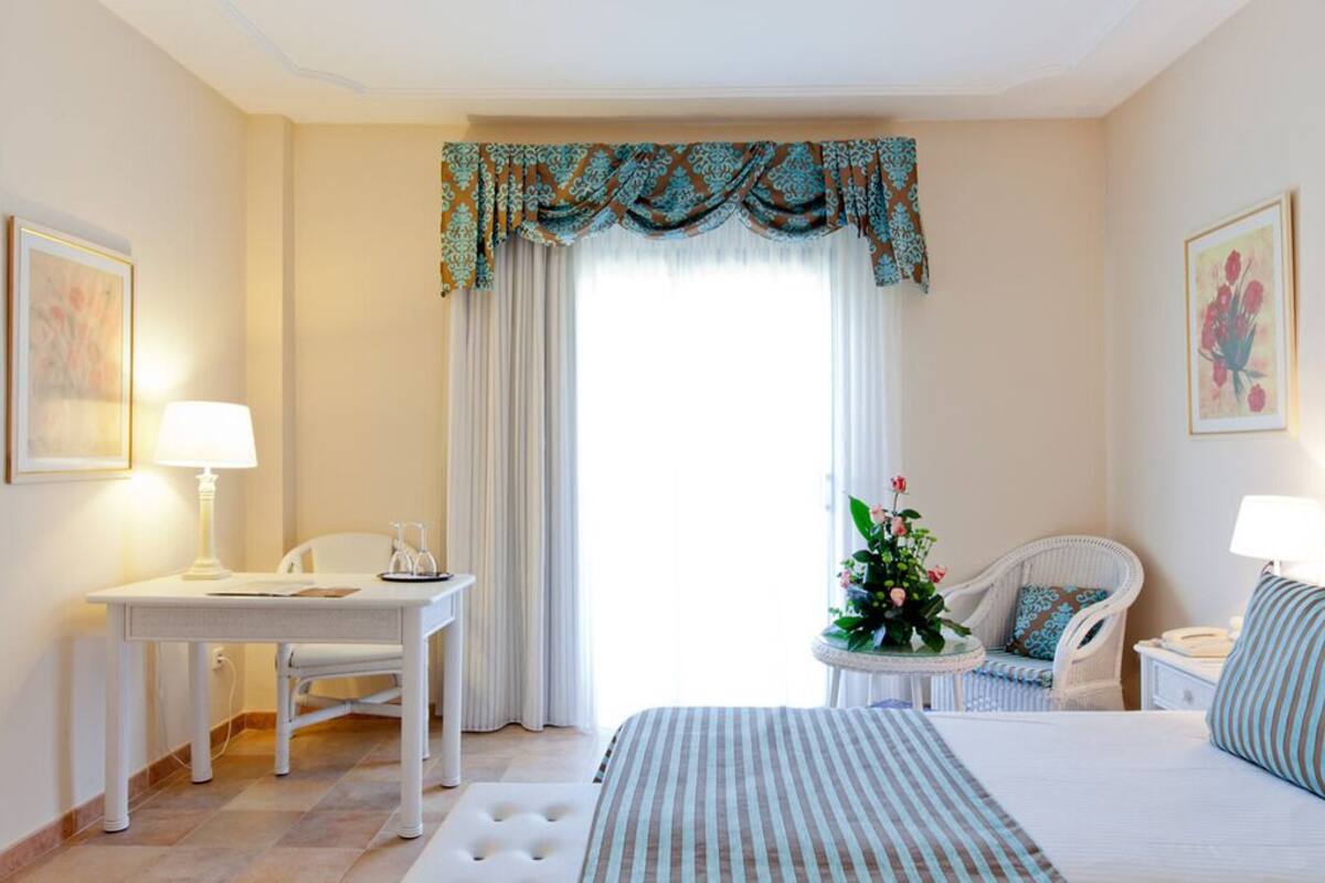 Habitación de hotel con cama, escritorio, dos sillas, una mesita con una planta y una ventana con cortinas.
