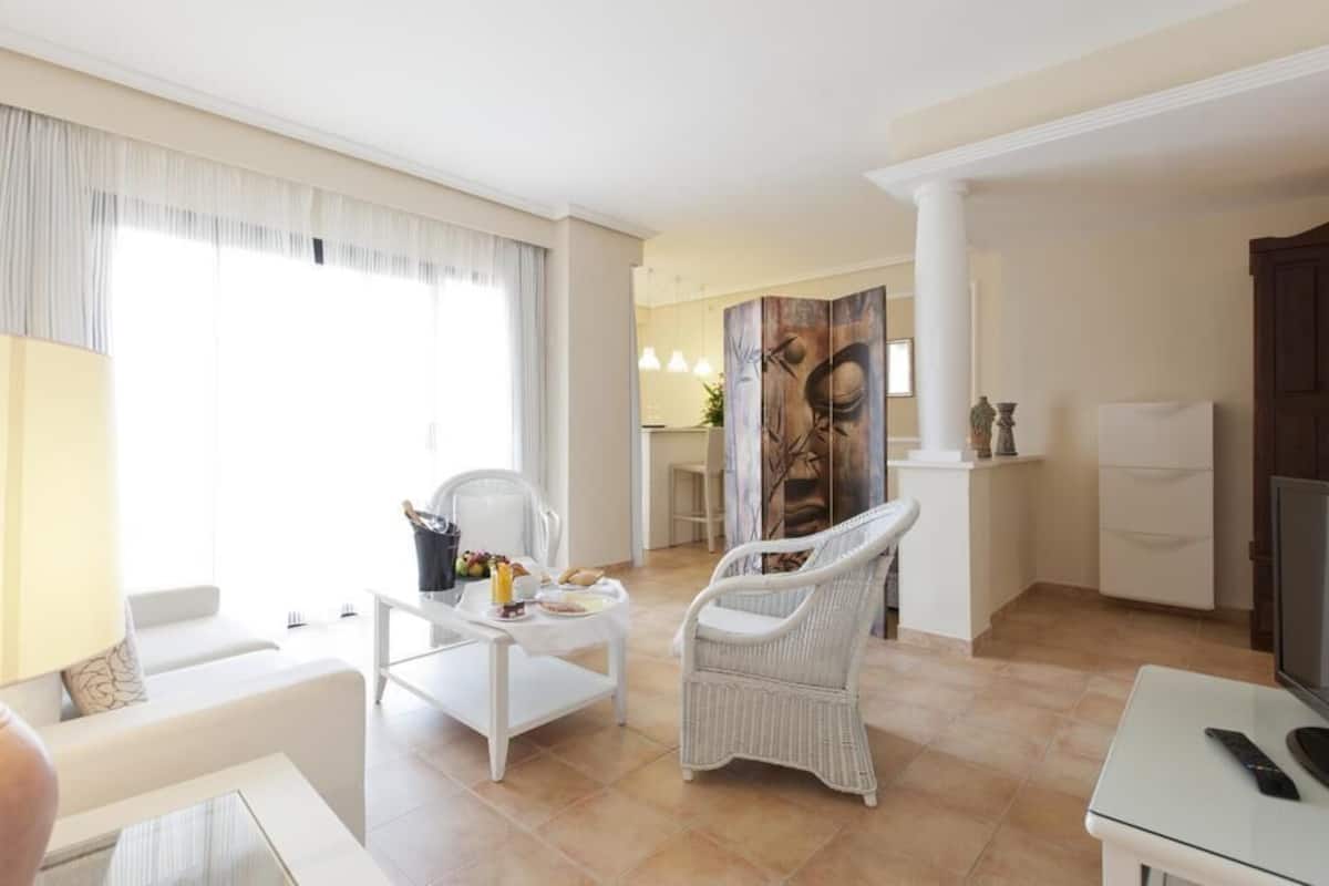 Un salón amplio con un sofá blanco, un sillón blanco, una mesa de centro y un televisor. Hay un cuadro en la pared y una ventana con cortinas.