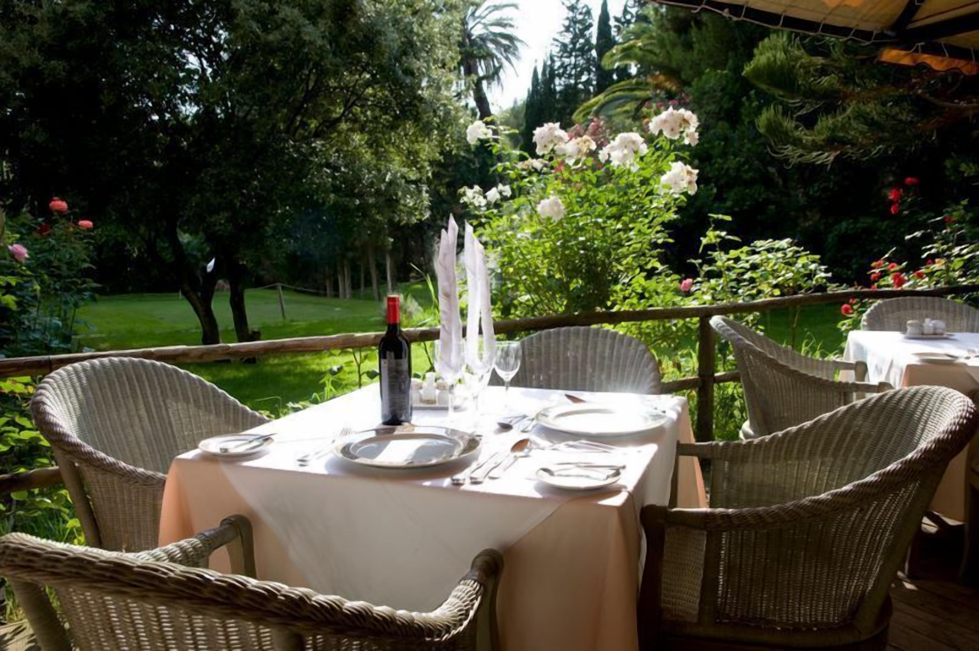 Un espace de restauration en plein air, avec des chaises en osier et une table dressée pour deux personnes, entouré d’une végétation luxuriante et de plantes en fleurs.