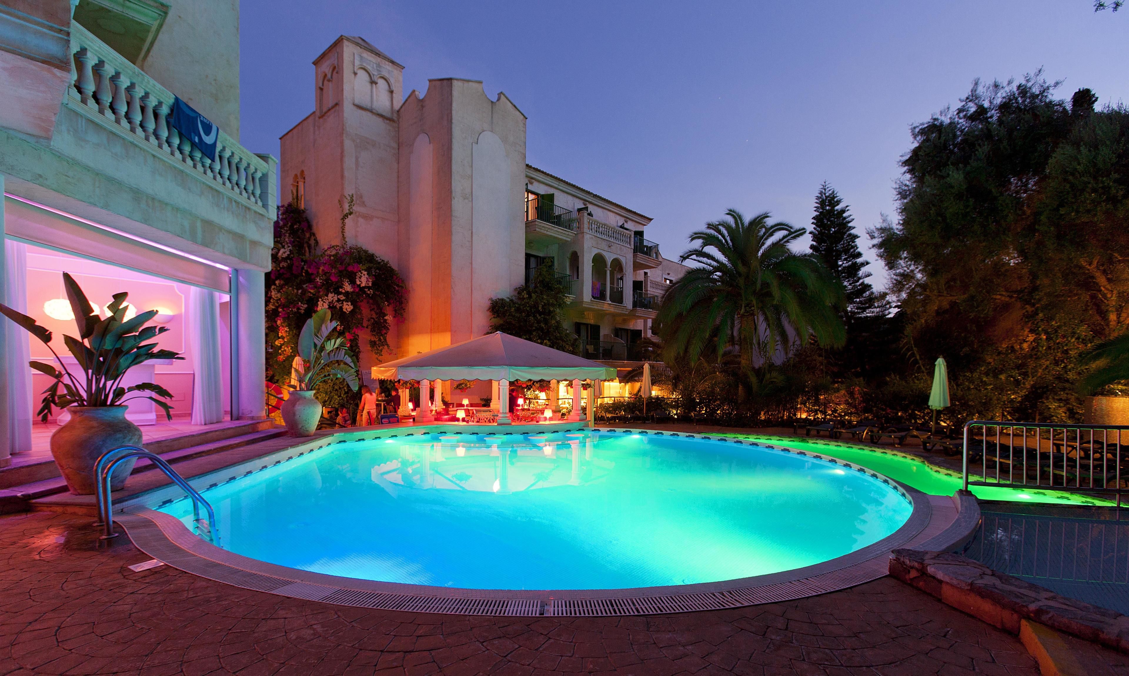 Zona de piscina con una fuente, rodeada de palmeras y un edificio con balcón.