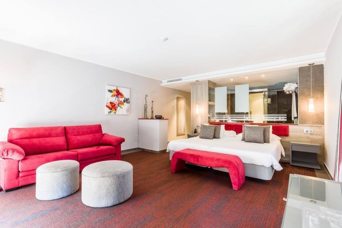 Una habitación de hotel moderna con un sofá rojo, una cama con ropa de cama roja y un espejo grande.