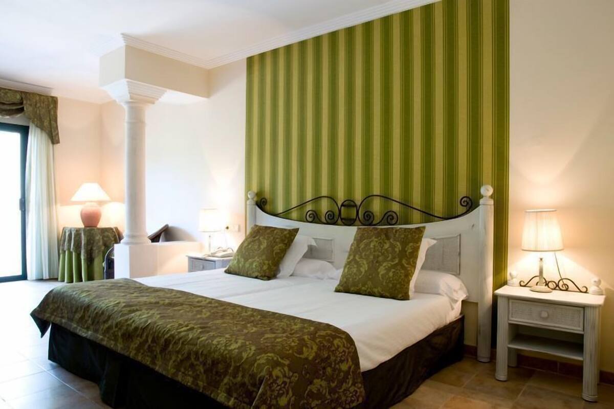Una cama bien hecha con un cabecero verde y blanco a rayas, dos mesitas de noche con lámparas y una ventana con cortinas.